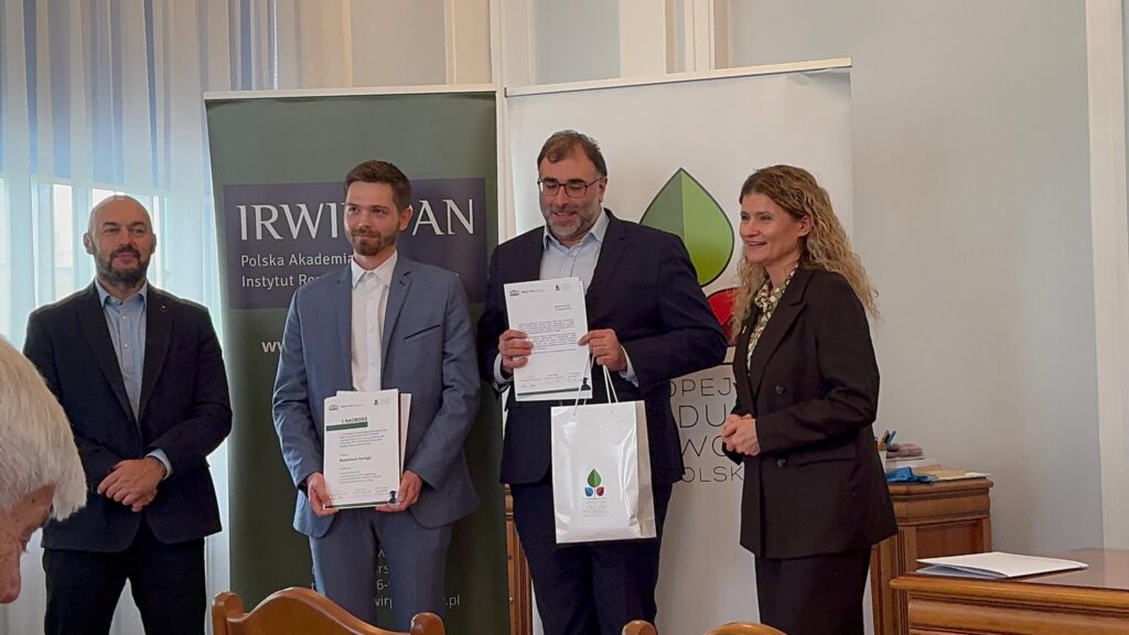 Zdjęcie przedstawia formalne wydarzenie odbywające się w pomieszczeniu, gdyż chodzi o ceremonię wręczenia nagród lub wyróżnień. W pierwszym planie stoją cztery osoby w eleganckich strojach — jedna z nich trzyma certyfikat i torbę z logo, a pozostali patrzą w jej stronę z uśmiechem. W tle widoczne są dwa banery z logo i nazwami „IRW” oraz „Polska Akademia Nauk”, co wskazuje na charakter naukowy lub instytucjonalny wydarzenia. Atmosfera jest uroczysta i profesjonalna, podkreślająca moment uznania lub osiągnięcia.