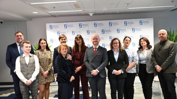 Zdjęcie przedstawia grupę dziesięciu osób, kobiet i mężczyzn, stojących blisko siebie w pomieszczeniu o charakterze konferencyjnym i pozujących do wspólnej fotografii; wszyscy są ubrani formalnie w stroje biznesowe, takie jak garnitury, marynarki, koszule i sukienki, a ich postawa jest profesjonalna i spokojna, z neutralnymi lub lekko uśmiechniętymi wyrazami twarzy. W tle widoczny jest duży biały baner z powtarzającymi się logotypami „Rzecznik Finansowy”, co wskazuje na oficjalne wydarzenie lub spotkanie związane z tą instytucją. Oświetlenie jest jasne i równomierne, typowe dla wnętrz, a całość tworzy wrażenie formalnej, uporządkowanej atmosfery charakterystycznej dla profesjonalnych wydarzeń lub konferencji.