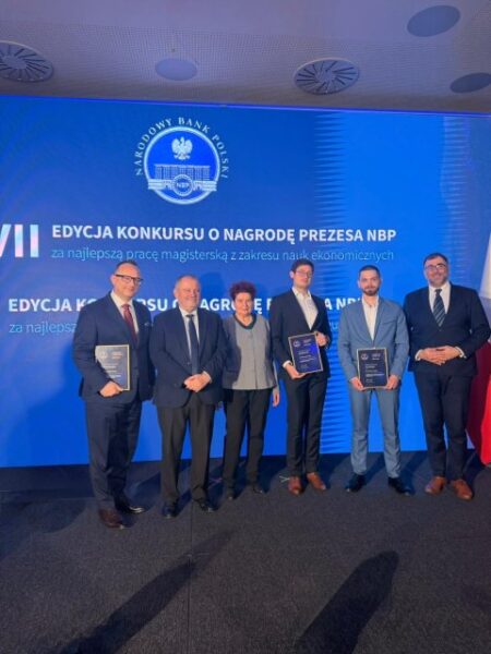 Obraz przedstawia formalne wydarzenie o charakterze uroczystym, podczas którego wyróżniane są osoby za osiągnięcia akademickie. Na pierwszym planie widać sześć osób stojących obok siebie na scenie, ubranych w eleganckie, formalne stroje, takie jak garnitury i krawaty. Trzymają one w dłoniach certyfikaty, co wskazuje na otrzymanie nagród lub oficjalnych wyróżnień. Postacie stoją w szeregu, zwrócone przodem do obiektywu, pozując do pamiątkowej fotografii, a ich postawa i wyraz twarzy sugerują profesjonalizm, dumę oraz poczucie spełnienia. W tle dominuje duży niebieski ekran z widocznymi logotypami i napisem „Narodowy Bank Polski”, a także informacją o konkursie o Nagrodę Prezydenta Narodowego Banku Polskiego za najlepszą pracę magisterską w dziedzinie nauk ekonomicznych. Sceneria wskazuje na wnętrze sali konferencyjnej lub audytorium, z wyraźnie zaznaczoną sceną i ciemną podłogą. Fotografia ma charakter reporterski i została wykonana podczas oficjalnej ceremonii, oddając atmosferę prestiżu, uznania oraz celebracji sukcesów naukowych.