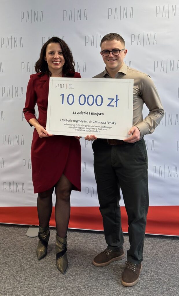 Dwie uśmiechnięte osoby stoją obok siebie na tle białej ścianki z powtarzającym się napisem „PA|NA”, trzymając wspólnie duży czek z informacją o nagrodzie 10 000 zł za zajęcie pierwszego miejsca i zdobycie nagrody im. dr. Zdzisława Fedaka. Osoba po lewej ma czerwony płaszcz i złote botki, a osoba po prawej beżową koszulę i okulary. Scena wygląda na zdjęcie z ceremonii wręczenia nagrody, wykonane we wnętrzu, na tle czerwonego dywanu.
