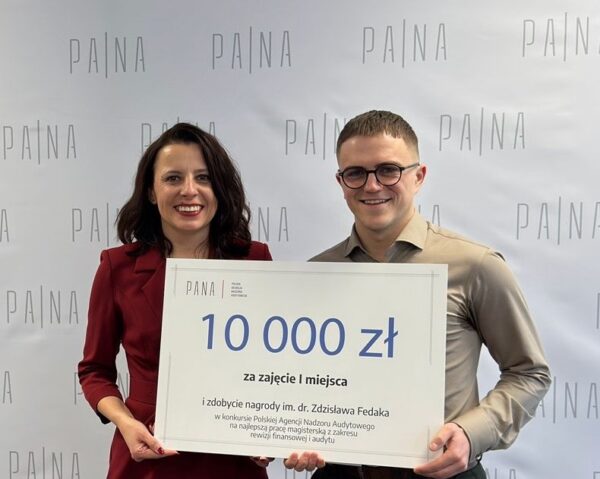 Dwie uśmiechnięte osoby stoją obok siebie na tle białej ścianki z powtarzającym się napisem „PA|NA”, trzymając wspólnie duży czek z informacją o nagrodzie 10 000 zł za zajęcie pierwszego miejsca i zdobycie nagrody im. dr. Zdzisława Fedaka. Osoba po lewej ma czerwony płaszcz i złote botki, a osoba po prawej beżową koszulę i okulary. Scena wygląda na zdjęcie z ceremonii wręczenia nagrody, wykonane we wnętrzu, na tle czerwonego dywanu.