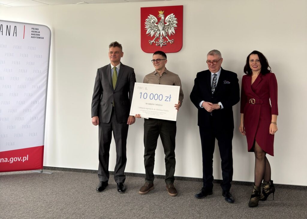 Cztery osoby w formalnych strojach stoją obok siebie na tle białej ściany podczas oficjalnego wydarzenia; druga od lewej trzyma duży czek z napisem „10 000 zł”. Po bokach stoją mężczyźni w garniturach, a po prawej kobieta w czerwonym żakiecie i czarnej sukni. W tle widoczny jest czerwony herb oraz baner z napisem „Polska Agencja Nadzoru Audytowego” i adresem „na.gov.pl”.