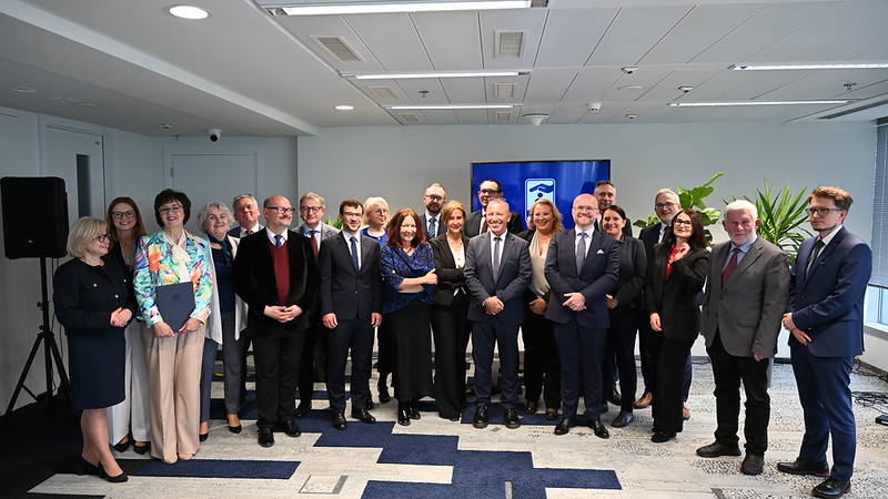 Grupa około 20 osób w strojach biznesowych pozuje do wspólnego zdjęcia podczas formalnego wydarzenia w nowoczesnym biurze lub sali konferencyjnej. Część uczestników trzyma dokumenty lub certyfikaty. W tle widoczny jest ekran z logo organizacji oraz rośliny dekoracyjne. Uczestnicy stoją w dwóch rzędach i patrzą w stronę aparatu.