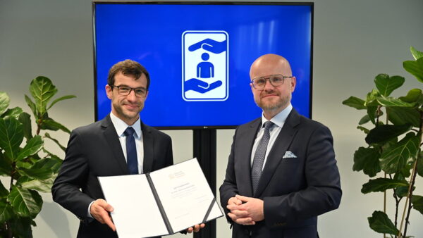 Dwóch mężczyzn w garniturach stoi przed ekranem z logo przedstawiającym dłonie otaczające sylwetkę człowieka. Mężczyzna po lewej trzyma otwartą teczkę z dokumentem. Zdjęcie wykonano podczas formalnego spotkania lub uroczystości w biurze albo sali konferencyjnej.